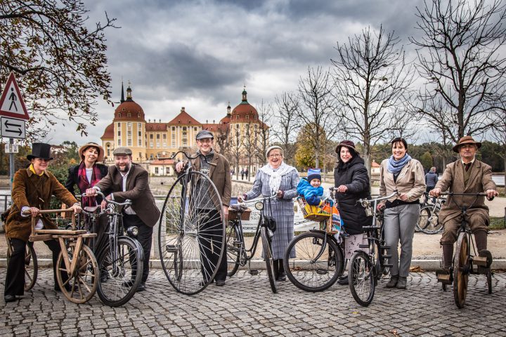 Vor dem Schloss Moritzburg sind in einer Reihe neun Frauen, Männer und Kinder mit ihren historischen Fahrrädern aufgereiht. Manche Personen tragen sogar historische Kleidung.
